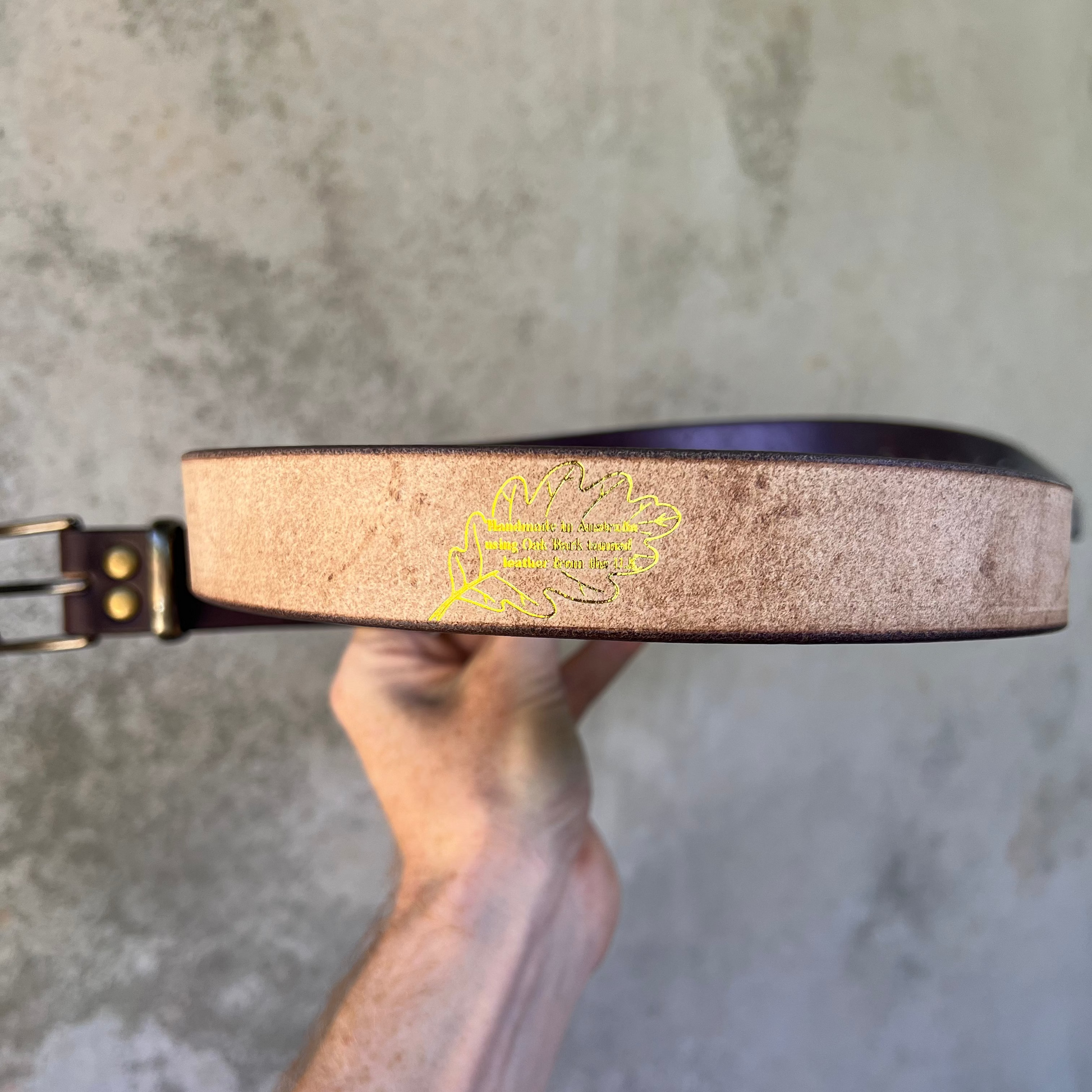 Hand holding a leather belt with a brand logo against a gray background