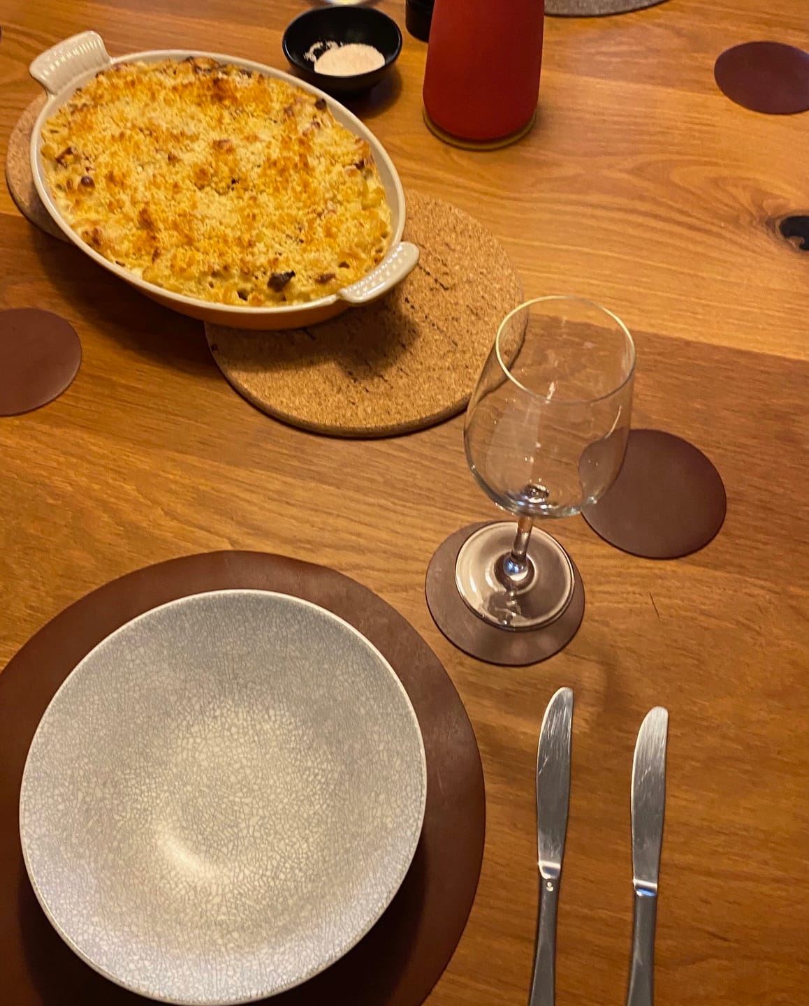 Dinner table setting with a dish of food, wine glass, and cutlery on a wooden surface.